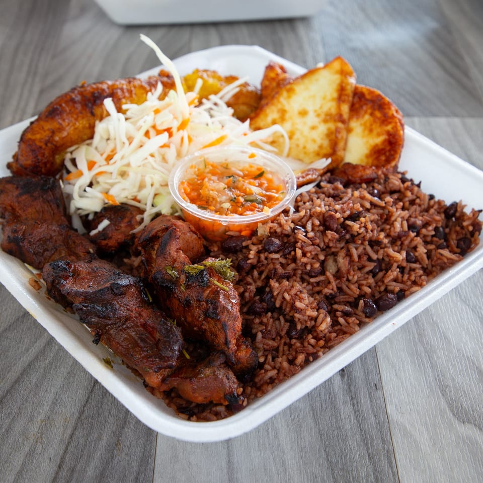 Takeout platter with grilled meat, red beans and rice, fried plantains, cabbage slaw, toasted bread, and spicy sauce.