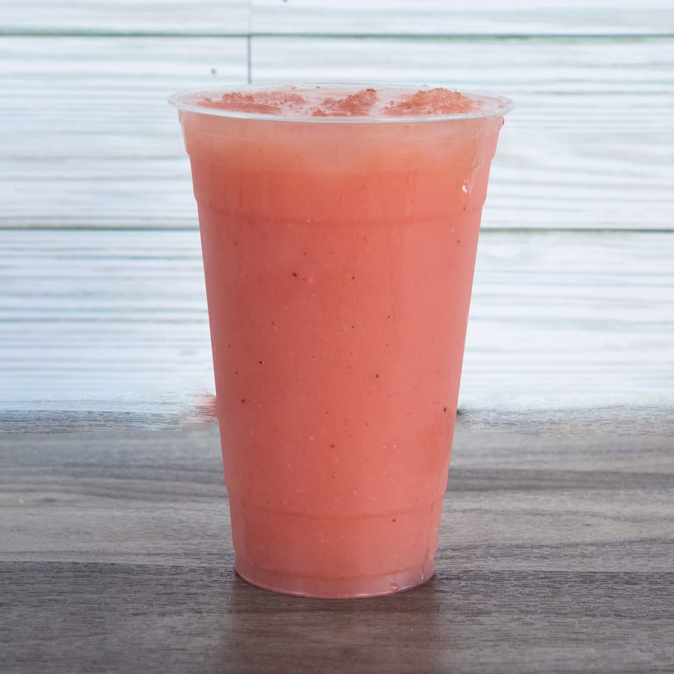 A clear plastic cup holds a frothy pink iced beverage on a wooden table against a white paneled wall.