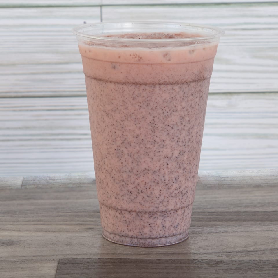 Clear plastic cup filled with a speckled pink smoothie on a wooden table against a white tiled wall.